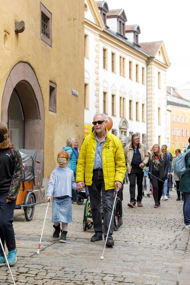 Fotografie: Ein blinder Mann mit Blindenstock führt ein Mädchen mit Augenbinde und Blindenstock durch die Neue-Waag-Gasse.