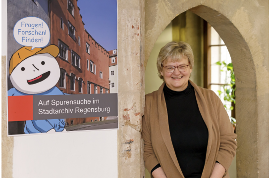 Fotografie: Martina Köglmeier lehnt an einer Mauer neben einem Plakat.