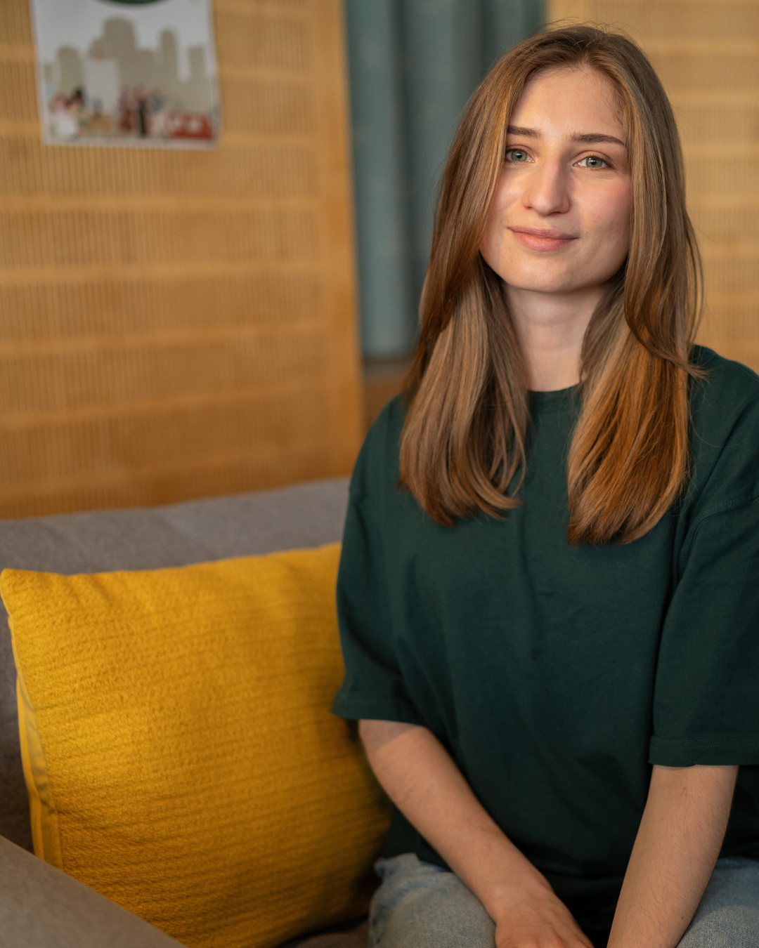 Auf dem Foto sieht man Maria Shevchenko, sie sitzt auf einer grauen Couch mit einem gelben Kissen und lächelt freundlich in die Kamera. Sie hat hellbraunes Haar und petrolfarbene Augen. Sie trägt ein ebenso petrolfarbenes Oberteil.