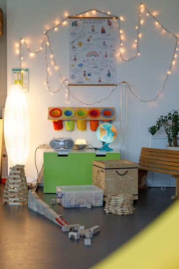 Fotografie: Spielzeugkisten, Bauklötze, ein CD-Player, ein Leuchtglobus, eine Holzbank, eine Zimmerpflanze, warm beleuchtet von einer Lichterkette und einer Papier-Stehlampe 