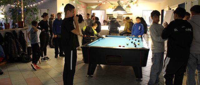 In der Mitte des Fotos ist ein blaubezogener Billardtisch zu sehen, um welchen zahlreiche Jugendliche stehen, während ein Billardspiel läuft. Im Hintergrund ist die Theke des Jugendzentrums zu sehen, an der auch einige Jugendliche sitzen.