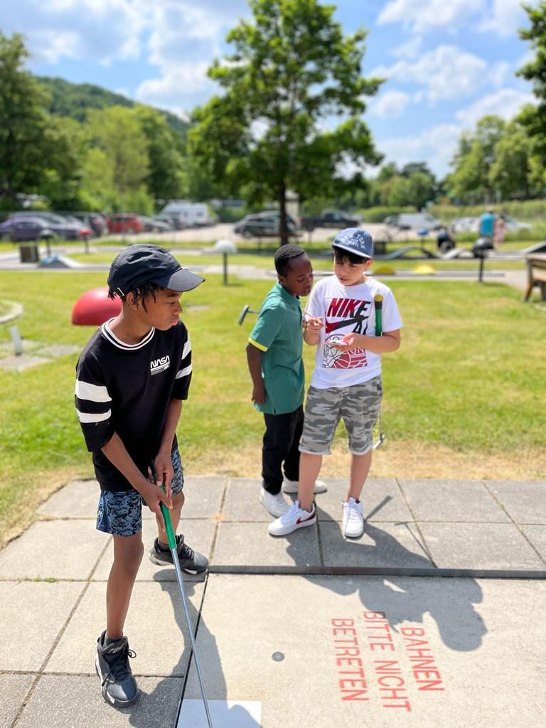 Auf dem Bild sind drei Kinder zu sehen. Zwei stehen neben der Minigolfbahn und betrachten etwas auf einem Smartphone während das dritte Kind gerade zum Schlag ausholt und Minigolf spielt.
