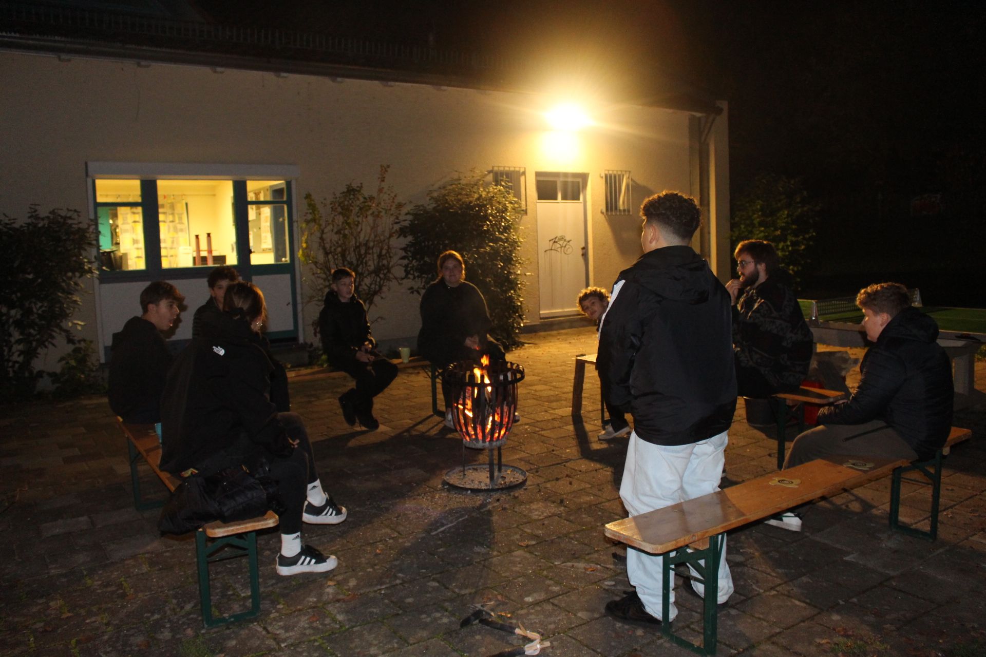 Das Bild zeigt eine Gesprächsrunde im Dunkeln auf dem Außengelände des Jugendzentrum. In der Mitte steht ein Feuerkorb um welchen in einem Viereck mehrere Bierbänke angeordnet sind, auf denen mehrere Menschen sitzen. Im Vordergrund des Bildes steht ein Kind, mit dem Rücken zu Kamera. Das Bild wird erhellt durch eine Außenleuchte am Haus.