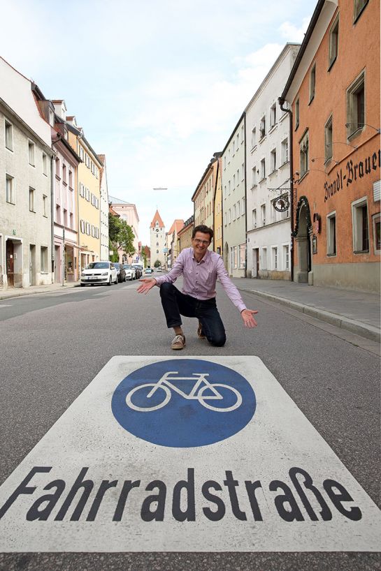 Fotografie: Im Vordergrund des Bildes befindet sich eine große Bodenmarkierung, die auf eine Fahrradstraße hinweist. Herr Großmüller kniet dahinter und präsentiert die Markierung. Das Foto wurde in der Ostengasse aufgenommen. 