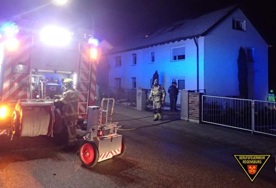 Fotografie: Feuerwehrfahrzeug vor einem Wohnhaus. Einsatzkräfte von Feuerwehr und Polizei im Hintergrund zu sehen.  