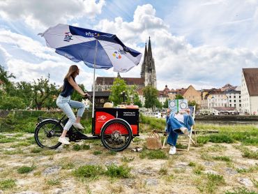 Fotografie – Eine Frau bringt auf dem Lastenrad das neue vhs Programm, eine Frau sitzt im Liegestuhl und schaut das Programm an. Im Hintergrund ist der Dom zu sehen