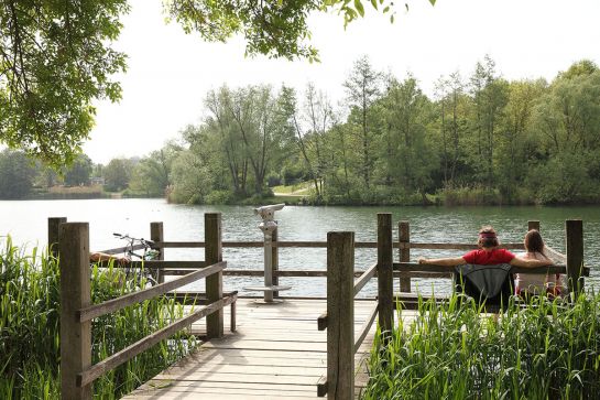 Fotografie - Steg im See, auf einer Bank sitzt ein Paar