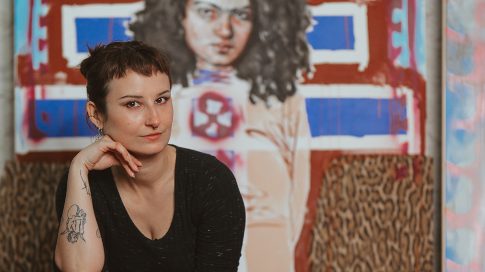 Die Abbildung zeigt die Künstlerin und Workshopleiterin Barbara Muhr sitzend auf einem Stuhl in ihrem Atelier vor einem ihrer Werke.