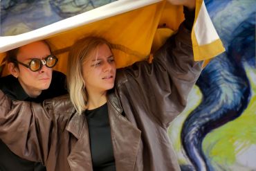 Fotografie von zwei Schauspielern, die sich unter einer Plane verstecken