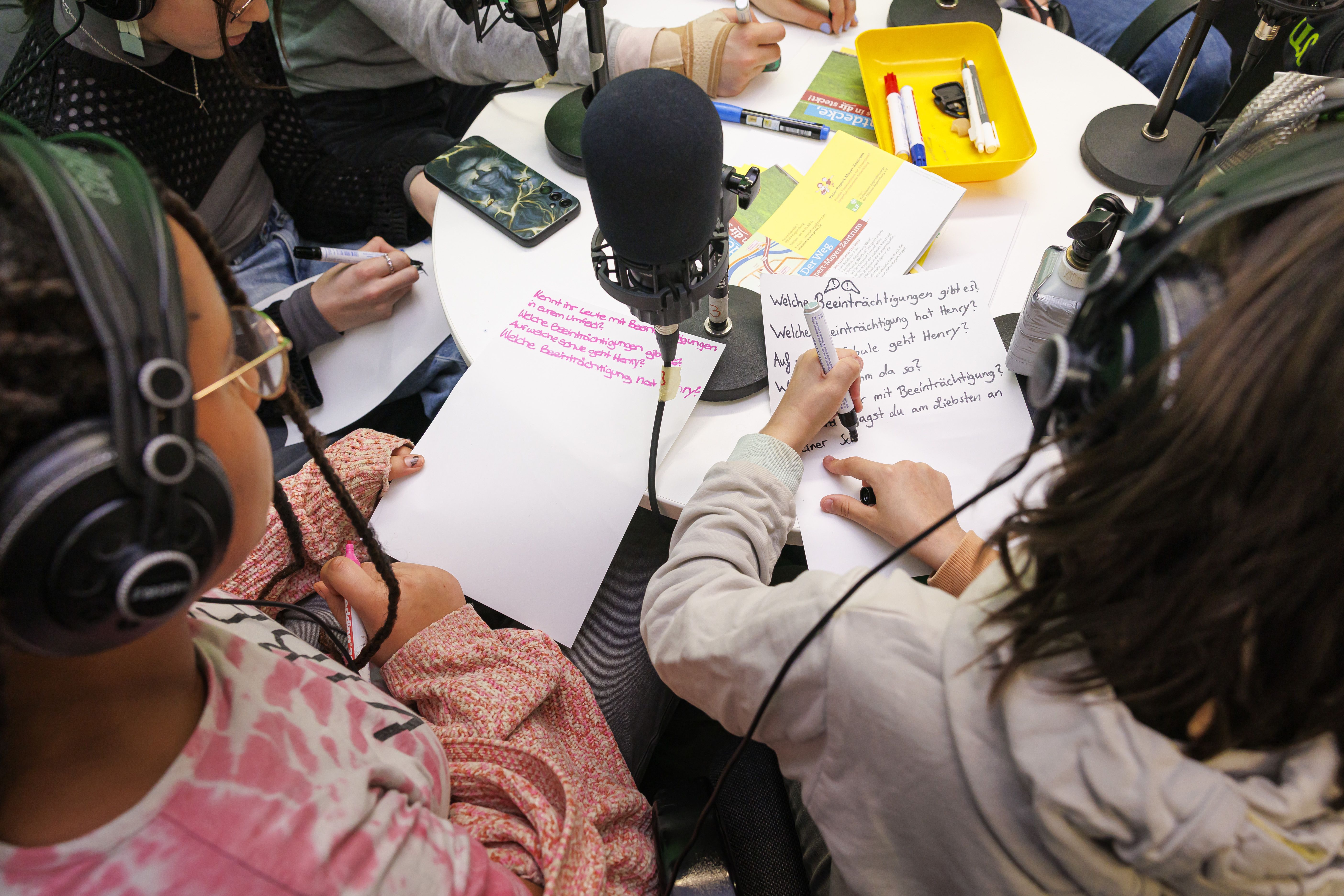 Aus der Vogelperspektive sieht man zwei Kinder, die vor einem Podcast-Mikrofon Zettel beschreiben.