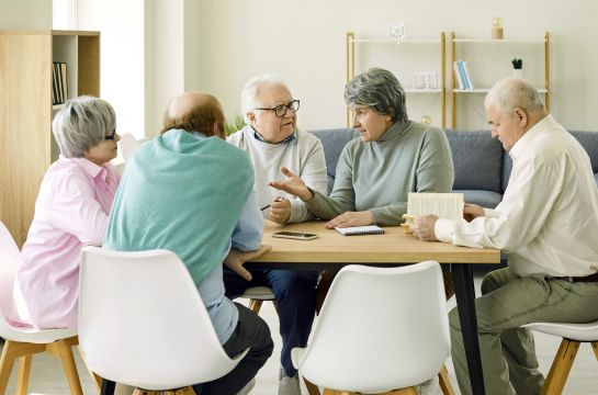 Fotografie: Fünf Seniorinnen und Senioren sitzen gemeinsam mit einem Buch am Tisch und reden.