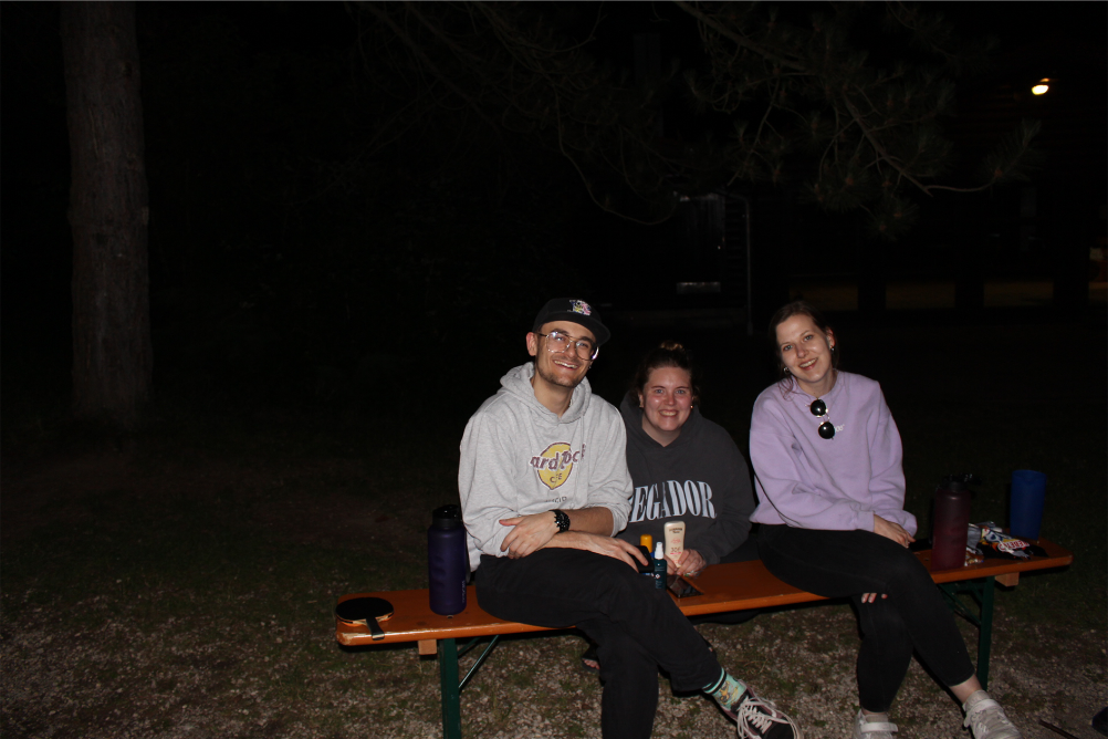 Drei Personen (zwei Frauen und ein Mann) sind abgebildet.  Eine Frau und ein Mann sitzen auf einer Bank, eine Frau sitzt in der Hocke hinter der Bank. Der Hintergrund ist dunkel und alle drei Personen blicken lachend in die Kamera.