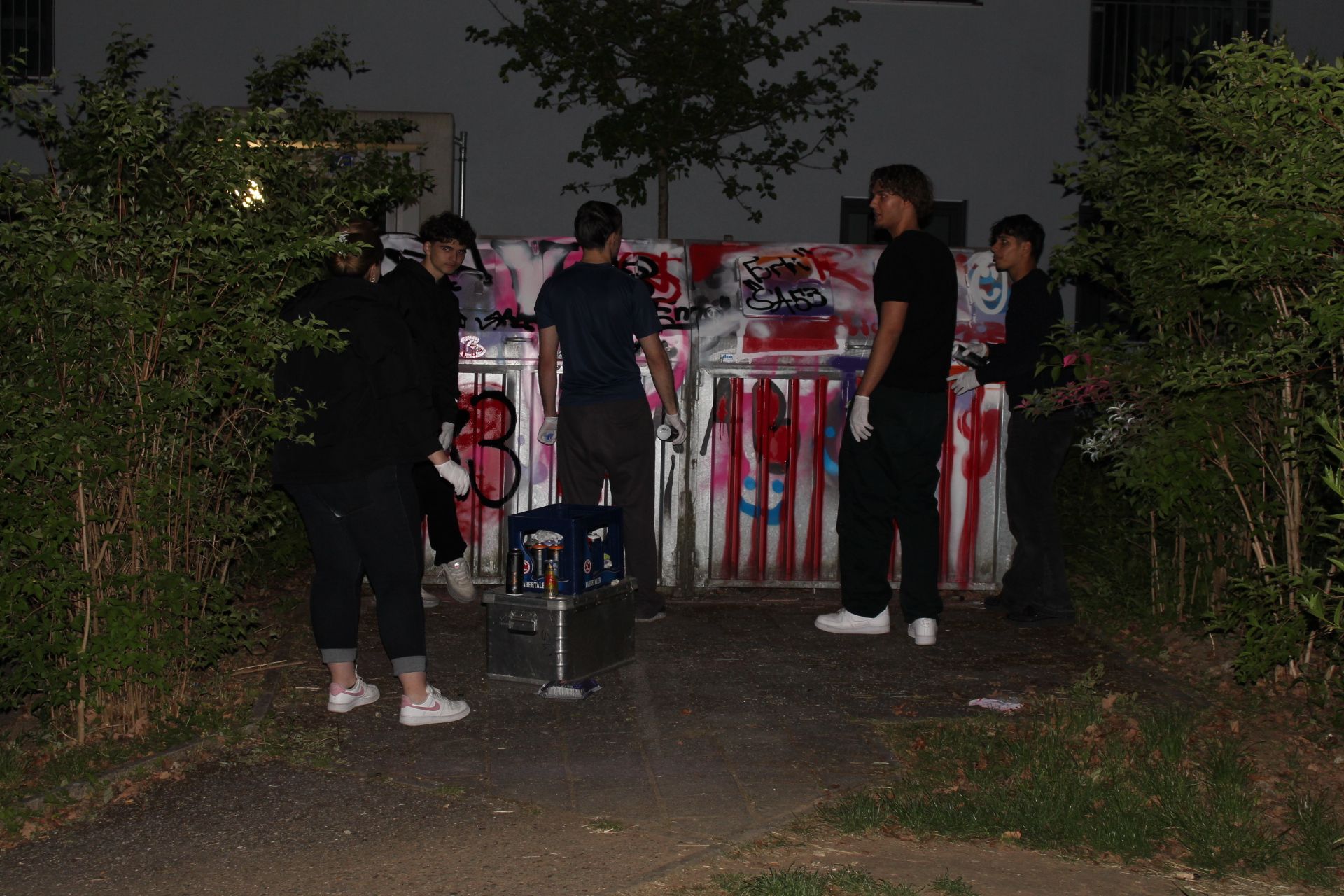 Auf dem Bild sind eine Mitarbeiterin des Jugendzentrums sowie vier Jugendliche zu sehen, die im Dunkeln die Mülltonnen des Jugendzentrums künstlerisch gestalten. Die Mülltonnen erstrahlen in bunten Farben - vorwiegend Pink, orange und weiß. Alle Beteiligten tragen Handschuhe und dunkle Kleidung.