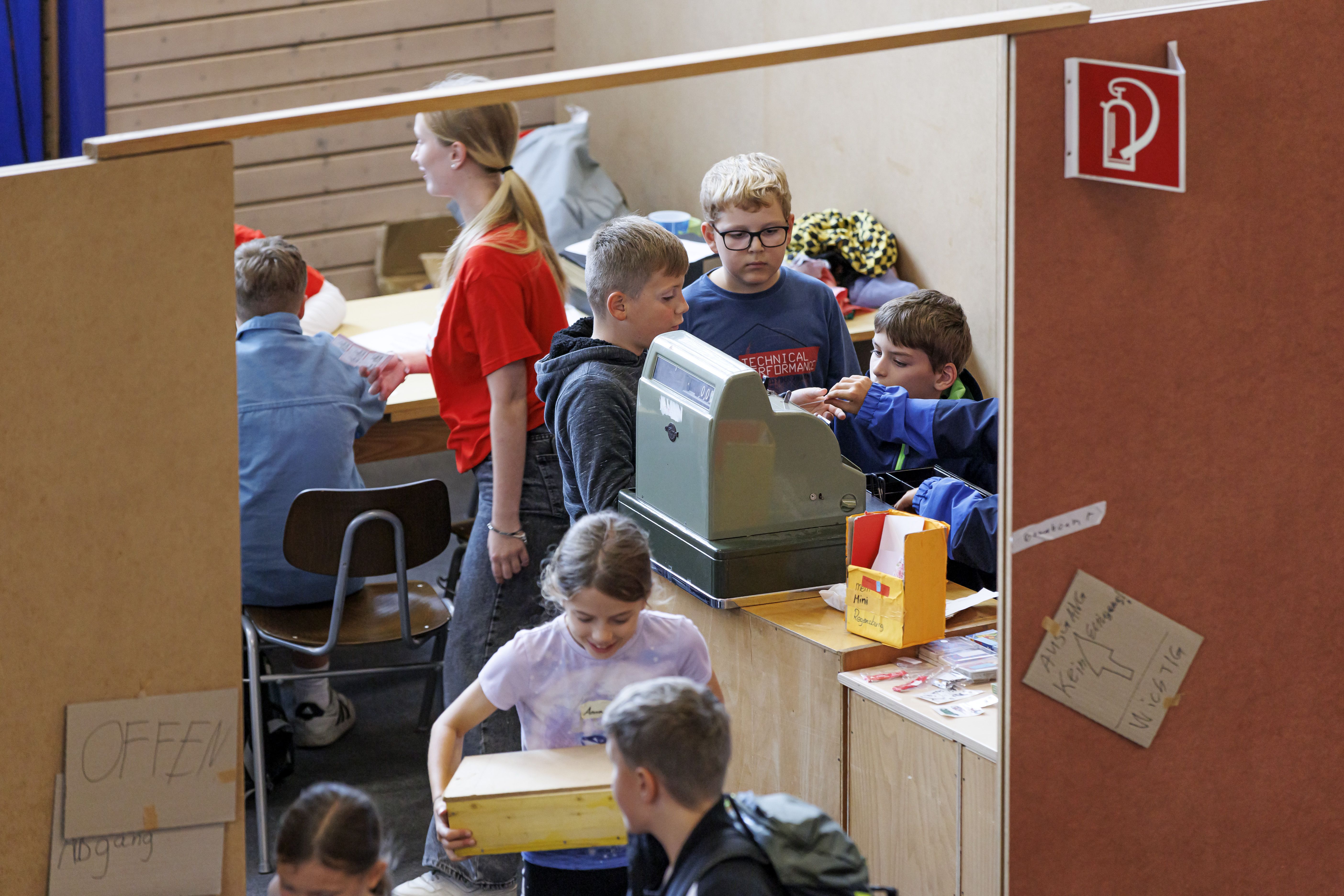 Fotografie: Blick ins Kaufhaus von Mini-Regensburg: Drei Kinder stehen um eine Registrierkasse, ein Mädchen trägt eine Holzkiste, im Hintergrund sind eine Betreuerin und weitere Kinder zu sehen