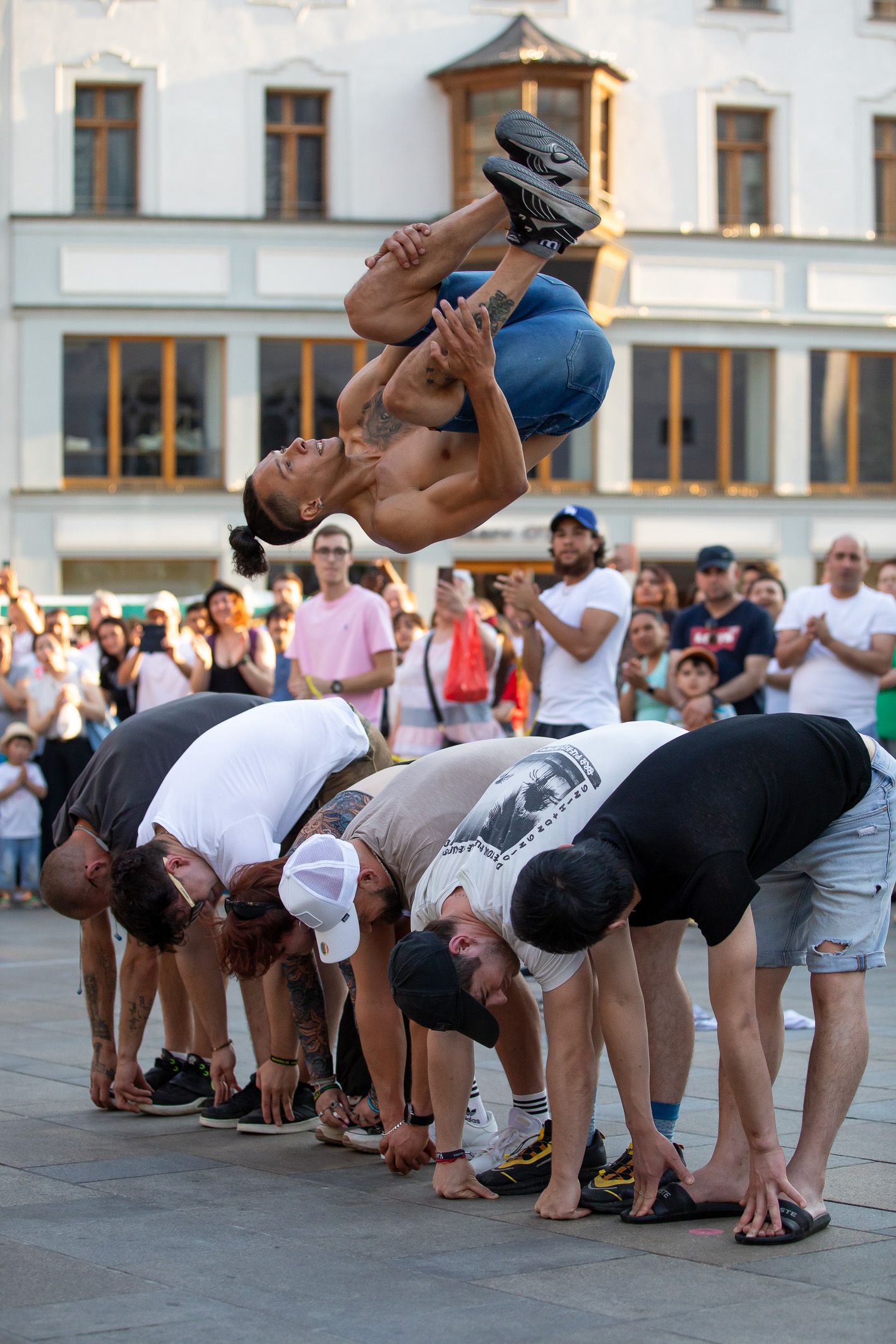 Fotografie: Ein junger Mann mit freiem Oberkörper macht einen Salto über sechs gebückte Menschen.