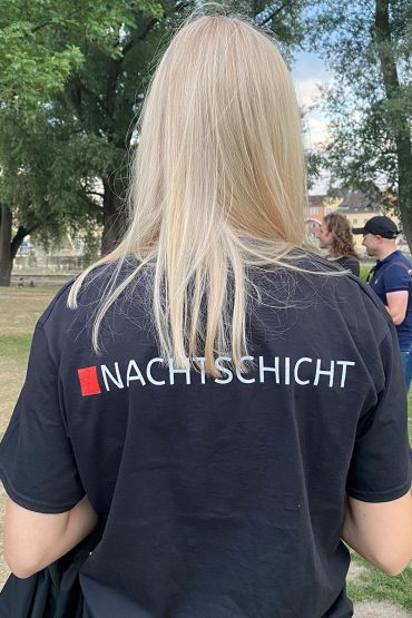 Fotografie - junger Mensch mit dem Rücken zur Kamera mit einem dunkelblauen T-Shirt, auf dem „Nachtschicht“ steht