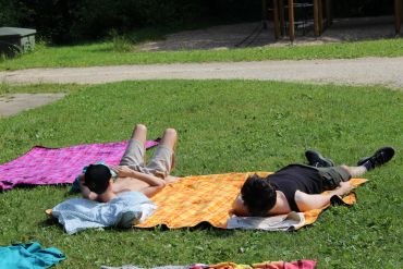 Auf dem Bild sind viele bunte Decken und Handtücher auf einer Wiese zu sehen. Links und rechts liegt jeweils ein Jugendlicher mit dunkler Kleidung auf den Decken und genießt die Sonne.