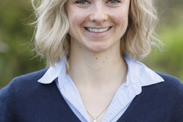 Man erkennt eine Frau mit blonden, kinnlangen, gewellten Haaren, die in die Kamera lächelt und ein blaues Hemd und darüber einen dunkelblauen Pullover trägt.