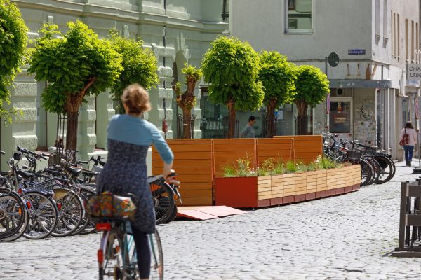 Fotografie: Eine Frau fährt auf einem Fahrrad vorbei an Stadtmöblierung und Bäumen.