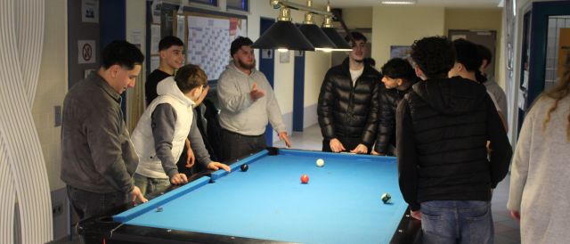 Im Zentrum des Bildes ist ein blaubezogener Billardtisch. Rundherum stehen einige junge Männer und Jugendliche, die sich angeregt unterhalten. Auf dem Tisch liegen ein paar Billardkugeln, da aktuell ein Spiel zugange ist.