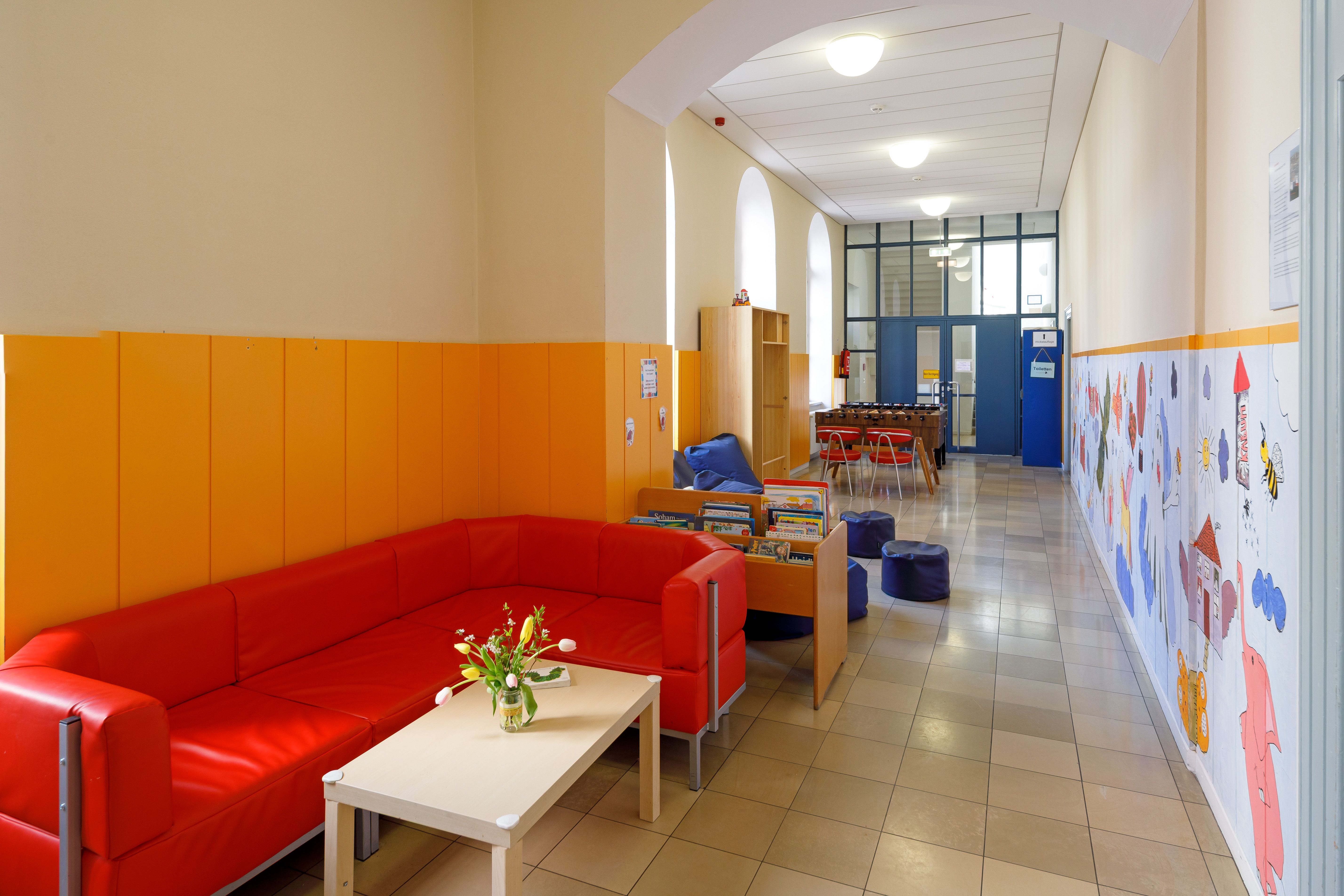 Langer, heller Flur mit von Kindern bemalter Wand. im Vordergrund ein gemütliches rotes Sofa, dahinter ein Regal mit Kinderbüchern, Sitzsäcke und Kicker