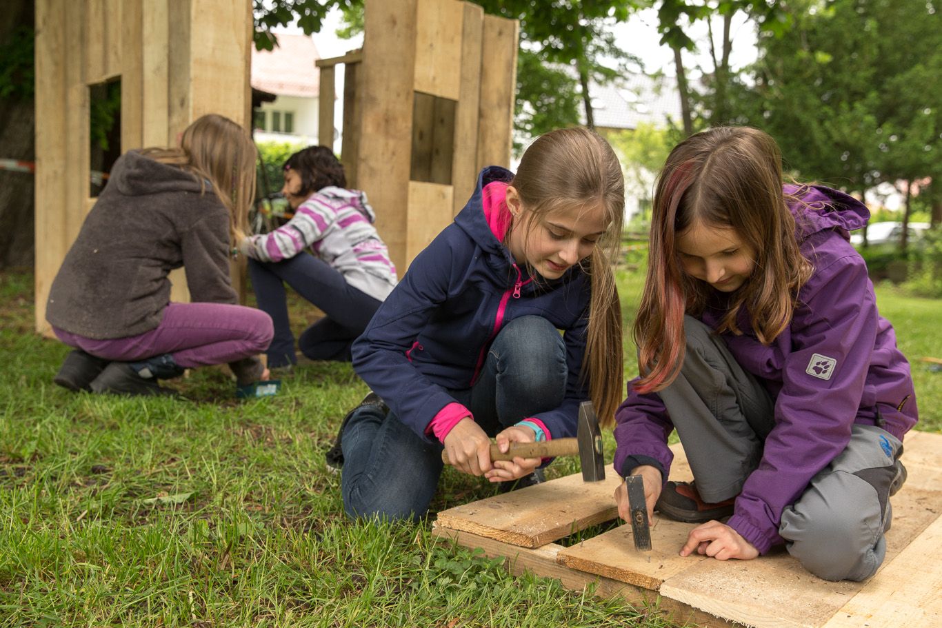 Fotografie – Mädchen bauen ein Holzhaus, dafür werden mit einem Hammer Nägel in am Boden liegende Bretter gehauen