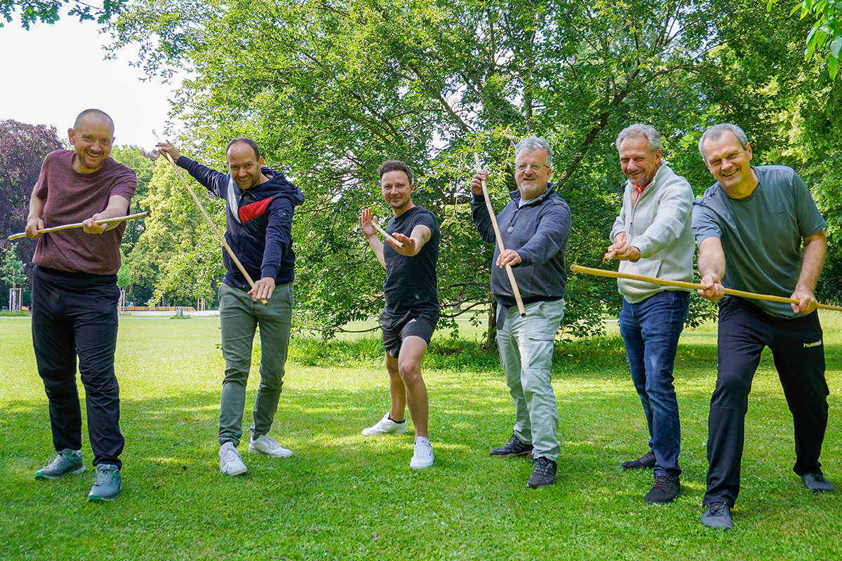 Fotografie - Sechs Männer trainieren mit Holzstöcken gemeinsam in einem Park.