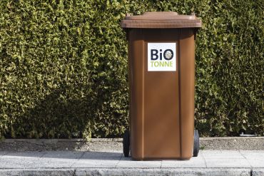 Fotografie: Eine braune Biotonnen steht vor einer Hecke
