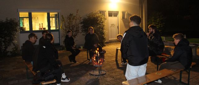 Das Bild zeigt eine Gesprächsrunde im Dunkeln auf dem Außengelände des Jugendzentrum. In der Mitte steht ein Feuerkorb um welchen in einem Viereck mehrere Bierbänke angeordnet sind, auf denen mehrere Menschen sitzen. Im Vordergrund des Bildes steht ein Kind, mit dem Rücken zu Kamera. Das Bild wird erhellt durch eine Außenleuchte am Haus.