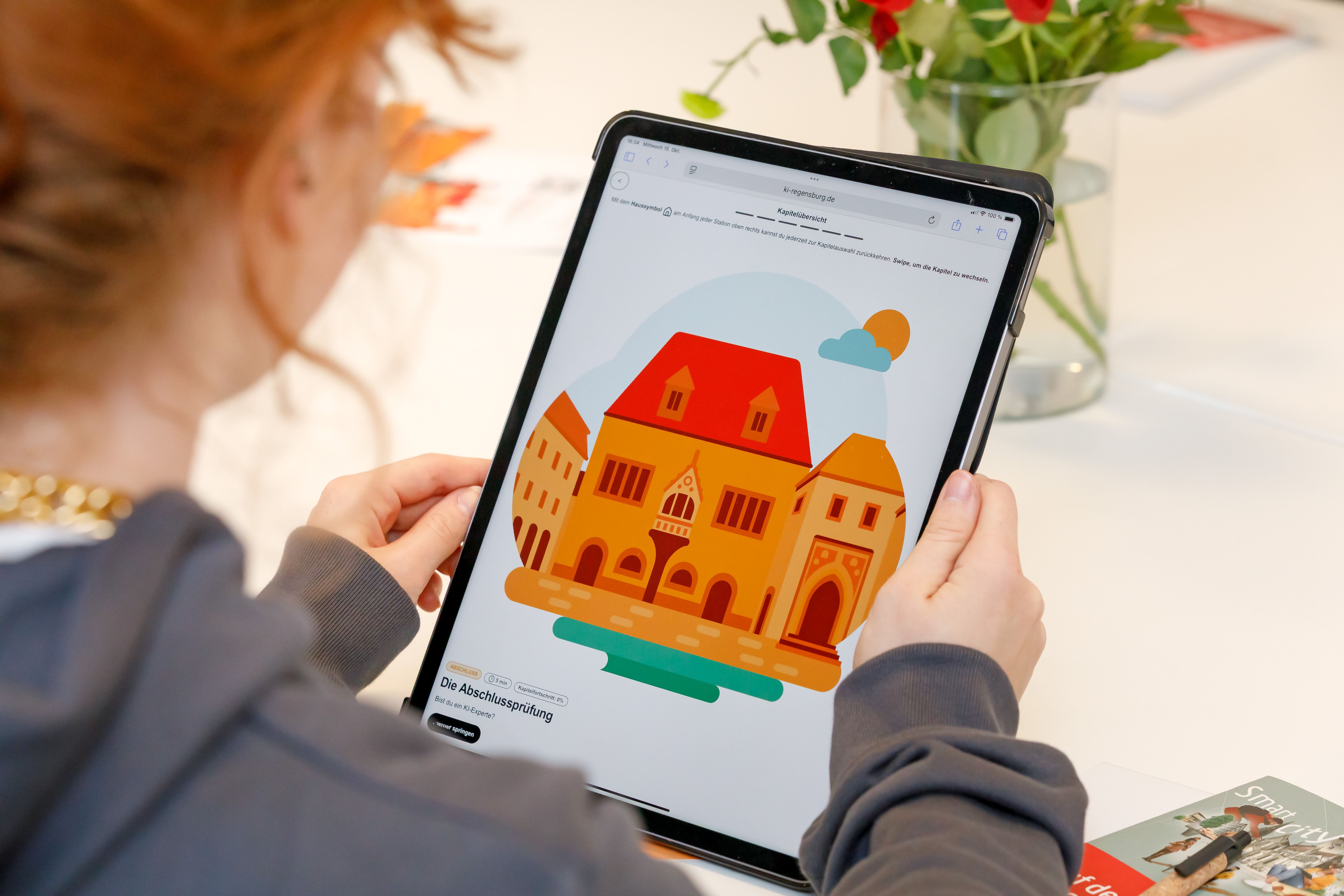Zu sehen ist ein Mädchen von hinten, das ein iPad in den Händen hält. auf dem Bildschirm ist die eine Seite des digitalen KI-Führerscheins zu sehen, auf der ein Rathaus abgebildet ist.