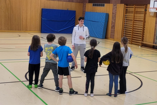 Fotografie -Jugendlicher erklärt einer Gruppe von Kindern ein Spiel in einer Sporthalle, einige halten Bälle.