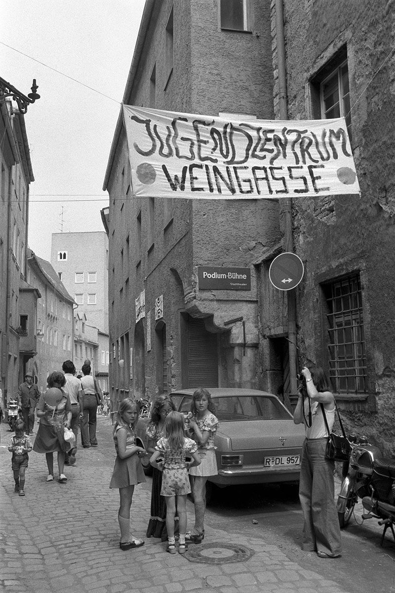 Zu sehen ist eine Aufnahme aus der Weingasse direkt vor dem neuen Jugendzentrum 1977. Quer über die Gasse wurde ein Banner mit der Aufschrift „Jugendzentrum Weingasse“ gespannt.                                    