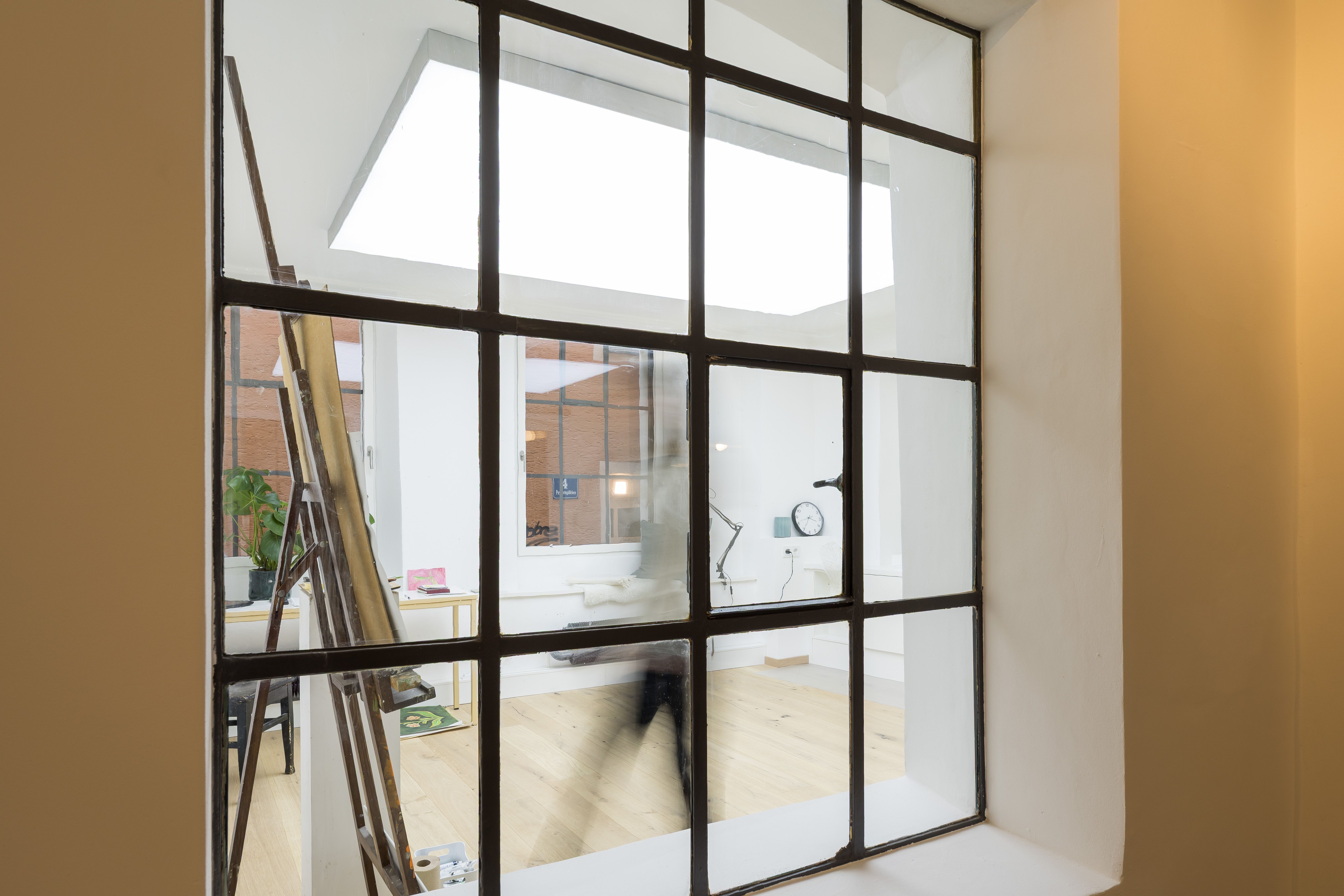 Fotografie - Atelierwohnung 1 - Der Blick geht durch ein für die Atelierwohnung charaktersistische Fenster mit Sprossenverteilung und zeigt das Studio mit einer verschwommenen weiblichen Silhouette, die durch das Bild läuft.
