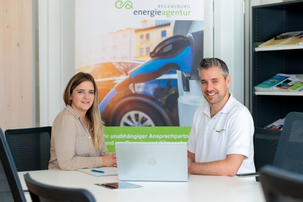 Im Bild sind eine Frau und ein Mann zu sehen, die an einem Tisch sitzen. Vor ihnen auf dem Tisch befindet sich ein aufgeklappter Laptop.
Im Hintergrund ist ein Plakat zu sehen, auf dem ein E-Auto beim Ladevorgang zu sehen ist. Weiterhin ist im Hintergrund ein Regal mit Broschüren zu sehen.