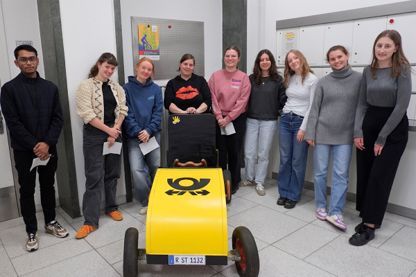 Fotografie - Gruppe von neun jungen Erwachsenen steht hinter einem gelben Postauto ähnlichen Seifenkistenfahrzeug in einem Gebäude.
