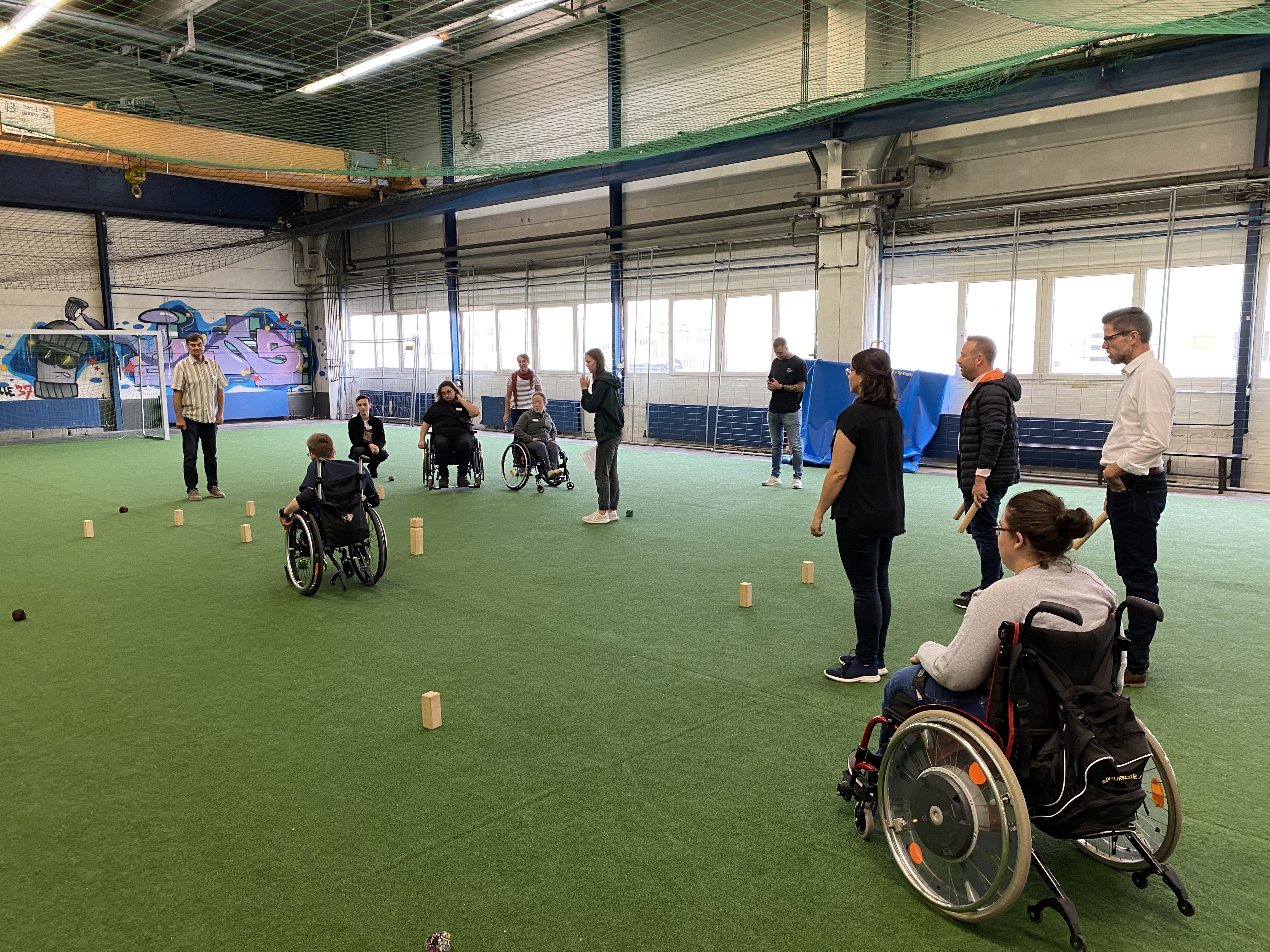 Fotografie: Mehrere Personen (darunter Personen im Rollstuhl) spielen ein Spiel in einer Halle.