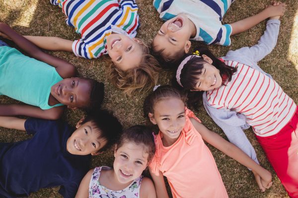 Das Bild zeigt eine diverse Kindergruppe. Die Kinder liegen am Boden im Kreis und halten sich an den Händen.