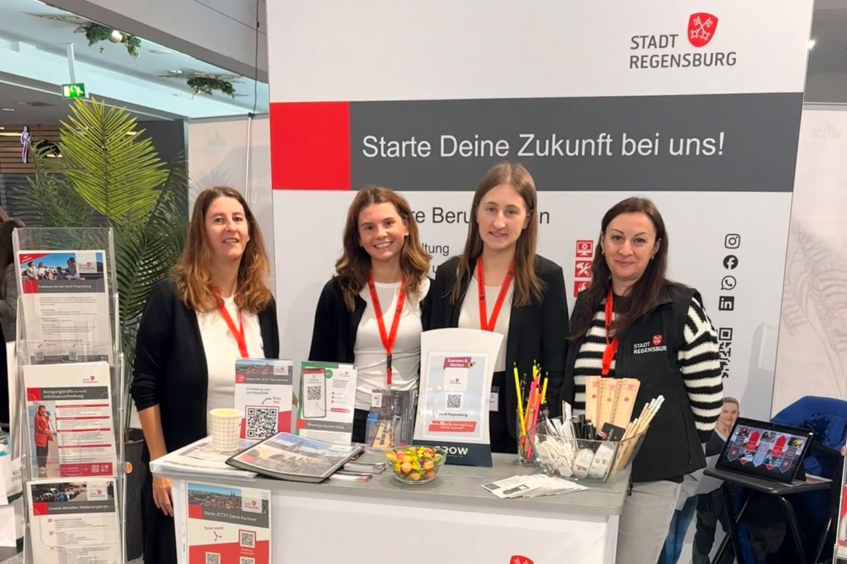 Mehrere Personen stehen an einem Messestand mit Informationsmaterialien und einem Roll-up-Banner; sie präsentieren das Ausbildungs- und Karriereangebot der Stadt Regensburg.