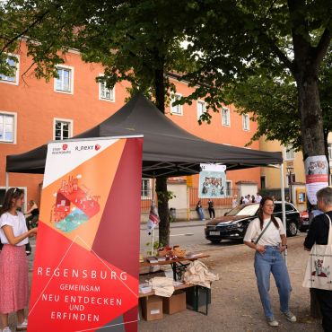 Im Vordergrund ist das R_NEXT-Roll-Up zu erkennen. Es trägt orangene und rote Dreiecke. Dahinter ist der Stand zu erkennen, an dem sich die Mitarbeitenden mit interessierten Besuchenden unterhalten.