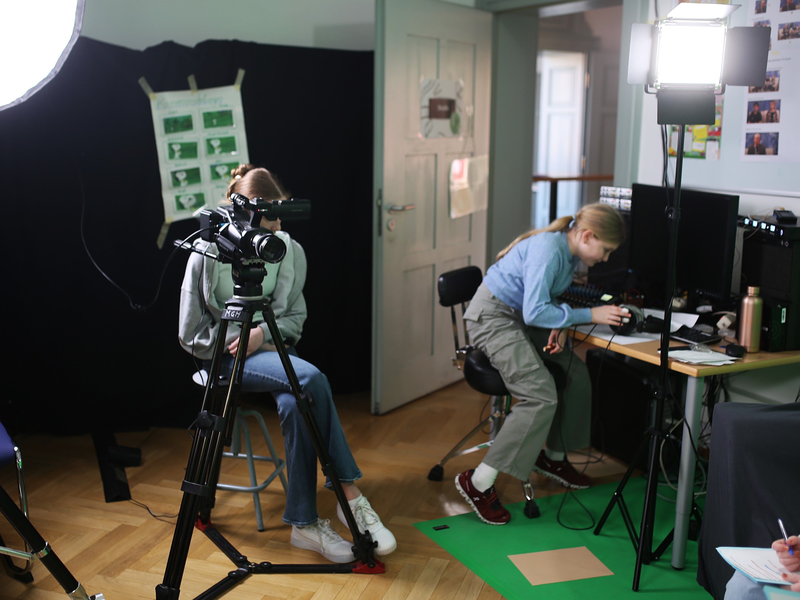 Links auf dem Bild sitzt ein Mädchen vor einer Filmkamera. Ihr Gesicht ist verdeckt. Rechts auf dem Bild sitzt ein Mädchen vor einem Mischpult und liest sich Notizen durch. Die Szenerie ist in einem Studio. Es sind Studiolichter und Greenscreen-Boden auszumachen.