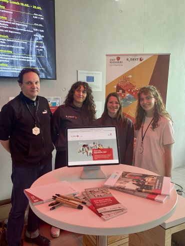 Ein Teil des R_NEXT-Teams steht hinter einem Messetisch, auf dem Infomaterial und auch ein iPad mit der R_NEXT-Website zu sehen ist. Im Hintergrund steht das Roll-Up von R_NEXT.