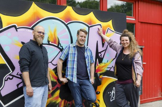 Das Team des Jugednzentrums (Benjamin Diestelmann, Lennart Krummeck und Esther Günther von links nach rechts) lehnt an einem Graffiti auf dem BOOM steht.