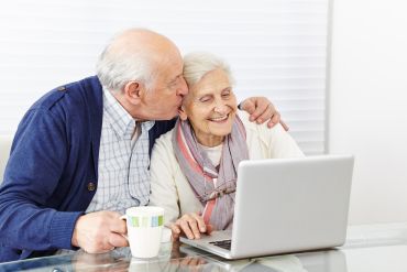 Fotografie: Ein Seniorenpaar sitzt vor einem Laptop. Der ältere Herr hat einen Arm um die Dame gelegt und gibt ihr ein Küsschen.