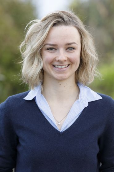 Man erkennt eine Frau mit blonden, kinnlangen, gewellten Haaren, die in die Kamera lächelt und ein blaues Hemd und darüber einen dunkelblauen Pullover trägt.