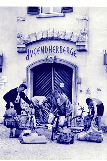 Zu sehen ist der Eingang der Wöhrdstraße 60 in Regensburg.  Über der doppelflügligen Eingangstür prangt die Hausnummer 60 und darüber der Schriftzug Jugendherberge. Auf der linken Seite neben der Tür sind an der Wand zwei Schilder zu erkennen auf denen „Stadtjugendamt -1. Stock-“ und „Erziehungsberatungsstelle“ Regensburg zu lesen ist. Im Vordergrund bepacken ein Mann und drei junge Erwachsene ihre Fahrräder mit Gepäck.