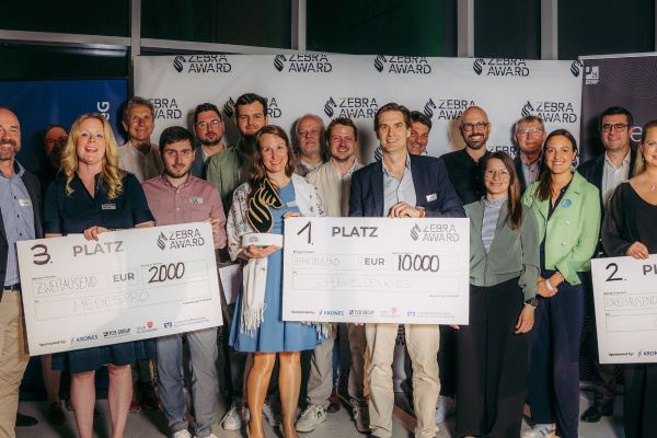 Eine Menschentraube steht vor einer Leinwand, auf dem das Motiv eines Zebras zu sehen ist sowie der Spruch "Zebra Award" drauf gedruckt wurde. Die Menschen in der ersten Reihe halten Preise von 2.000€, 3.000€ und 10.000€ in den Händen.