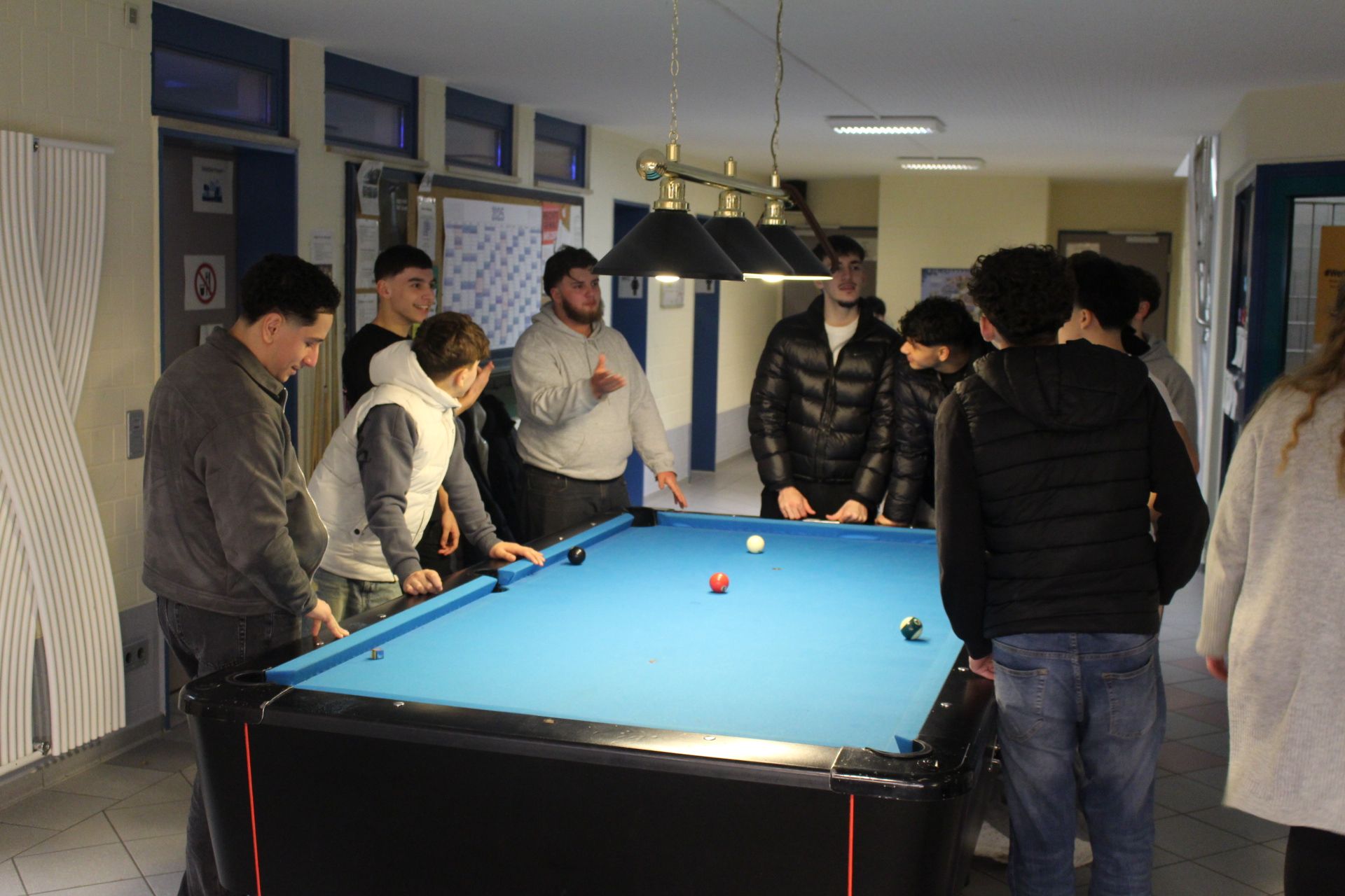 Im Zentrum des Bildes ist ein blaubezogener Billardtisch. Rundherum stehen einige junge Männer und Jugendliche, die sich angeregt unterhalten. Auf dem Tisch liegen ein paar Billardkugeln, da aktuell ein Spiel zugange ist.
