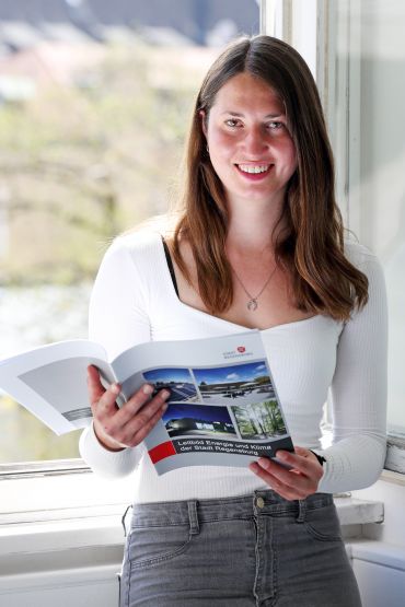 Fotografie: Verena Schmid lehnt am Fensterbrett und hält eine Broschüre in den händen.