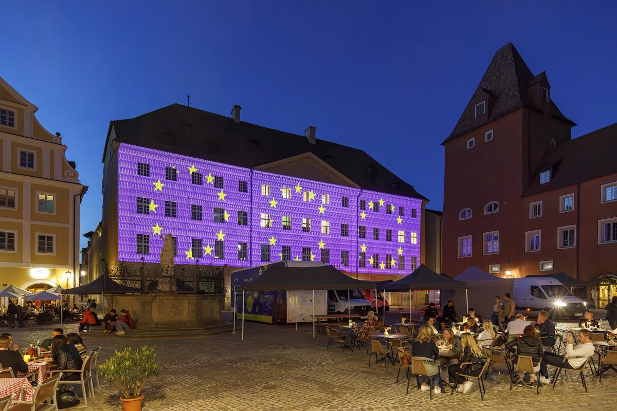 Am Haidplatz in der Regensburger Innenstadt werden auf die Fassade des Thon-Dittmer-Palais  EU-Motive mit blauem Hintergrund und gelben Sternen projiziert. Auf dem Platz sind rund um den Brunnen Pavillons zu sehen. Viele Personen nutzen das Angebot der Außengastronomie