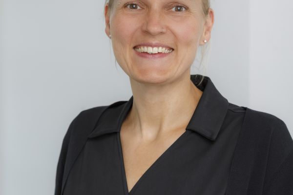 In der Mitte des Bildes steht eine breit lächelnde Frau mit blonden Haaren, die sie zu einem Dutt gebunden hat. Sie trägt Perlenohrringe und ein schwarzes Wickelkleid.